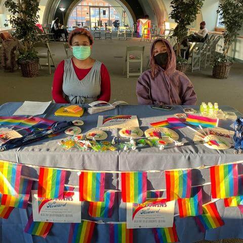 a picture of two students at a table at the entrance of the event. lots of rainbows